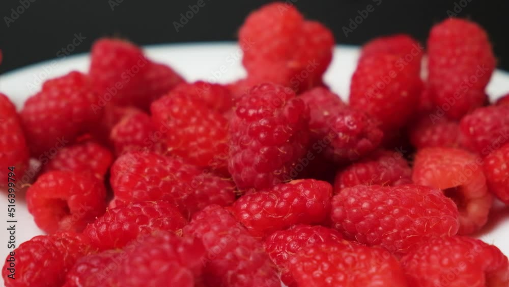 Raspberries fall into a white plate on a black background, side view, slow motion. Raspberries fall into a white plate on a black background, side view, slow motion.