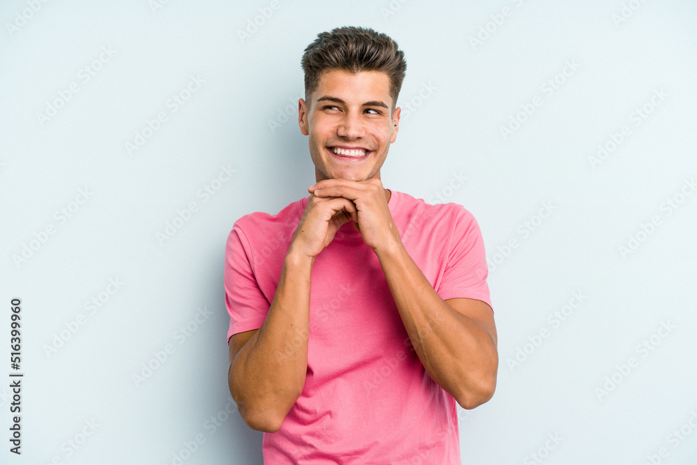 Young caucasian man isolated on blue background keeps hands under chin, is looking happily aside. Young caucasian man isolated on blue background keeps hands under chin, is looking happily aside.