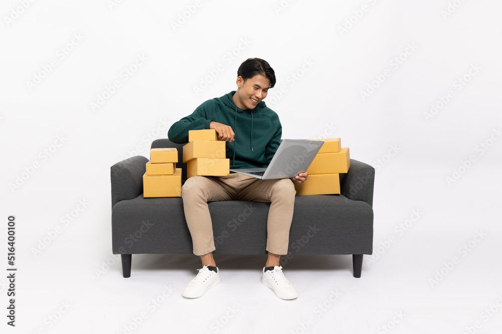 Young Asian man sitting on sofa with laptop computer and package parcel box isolated on white background, Delivery courier and shipping service concept