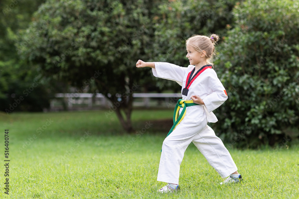 Caucasian little girl seven years old in kimono with yellow green belt exercising Taekwondo at summer park alone during coronavirus covid-19 lockdown, self isolation and social distancing Caucasian little girl seven years old in kimono with yellow green belt exercising Taekwondo at summer park alone during coronavirus covid-19 lockdown, self isolation and social distancing