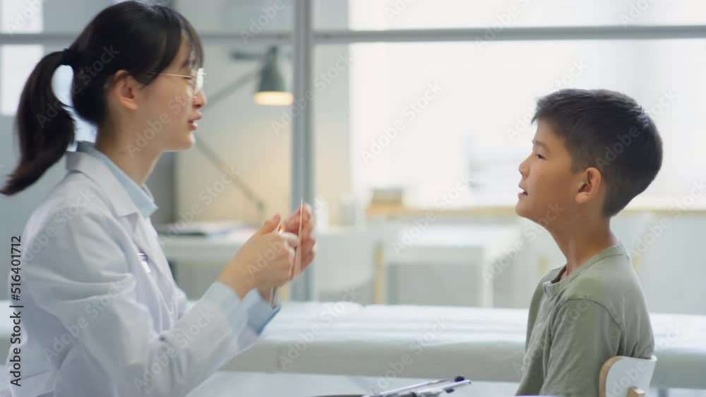 Asian female doctor examining throat of tween boy with flashlight during medical checkup in clinic Asian female doctor examining throat of tween boy with flashlight during medical checkup in clinic