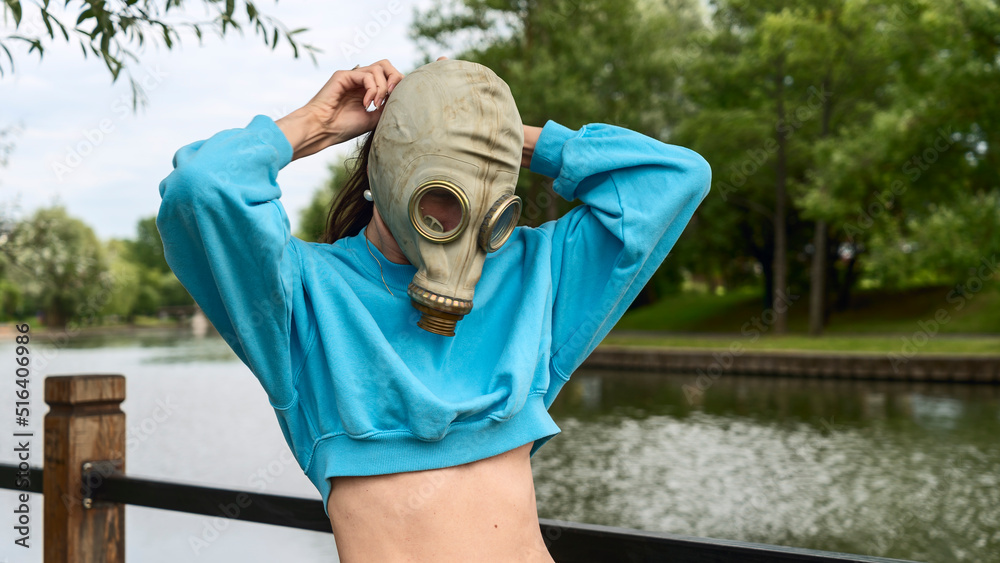 Naklejka premium The girl puts on a gas mask on the river bank. Alienation in the city. Fashion, ecology and loneliness concepts.