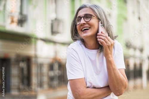 Charming mature business woman with grey hair talking on the phone holding smartphone standing outdoors of the streets of old urban city wearing white t-shirt. Silver hair woman outdoors