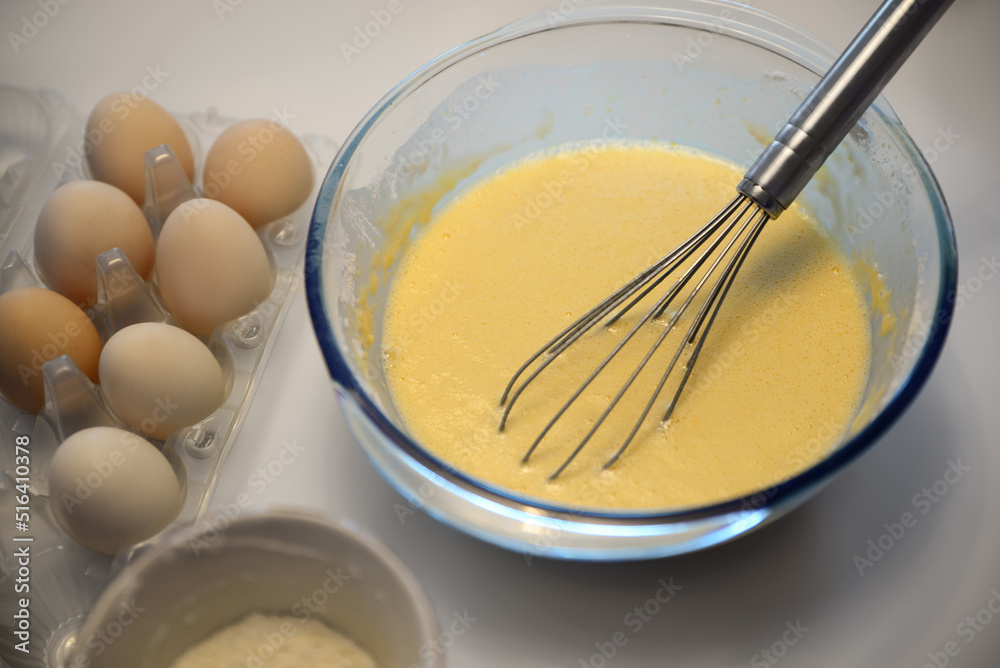 Glass bowl of crepe batter with whisk and ingredients on white table, closeup Glass bowl of crepe batter with whisk and ingredients on white table, closeup