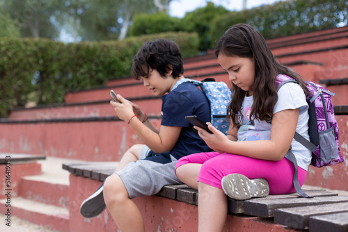 Caucasian boy and girl primary school students using mobile phones. They are outdoors. Concept of using technology at children's age.