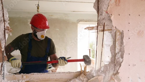 Contractor wrecks wall with sledgehammer making hole for rearrangement. Man doing manual dismantling and demolition works with big hammer hits for apartment renovation. Construction worker in uniform.