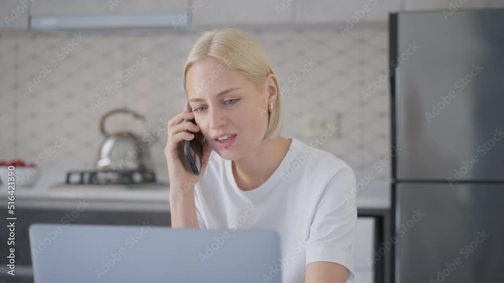 Attractive blonde woman talks on phone while working on laptop in kitchen Attractive blonde woman talks on phone while working on laptop in kitchen