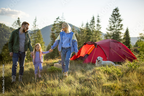 Young caucasian family with little girl walk together on green meadow while traveling with tent in the mountains. Happy family summer vacation at campsite