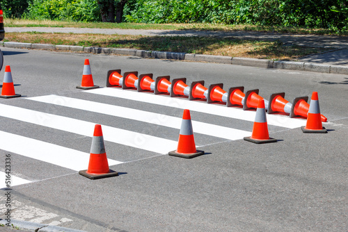 A newly painted pedestrian crossing across the road is fenced off with traffic cones on a sunny summer day.