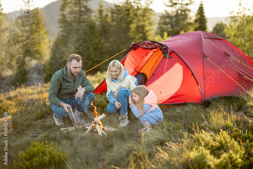 Young adult couple sit with a little girl by the campfire at campsite, traveling with tent on nature. Family spend summer time hiking in the mountains