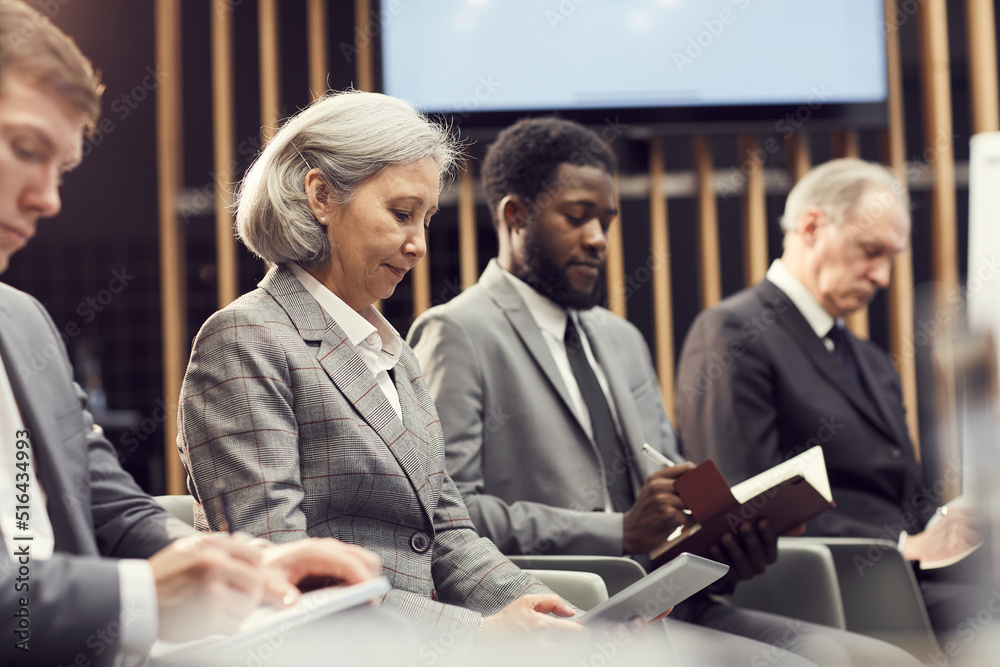 Group of serious multi-ethnic business people sitting in row and making notes while summarizing information at conference Group of serious multi-ethnic business people sitting in row and making notes while summarizing information at conference