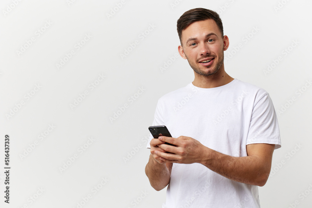 Cheerful tanned handsome man in basic t-shirt typing message with phone smile at camera posing isolated on over white studio background. Copy space Banner Mockup. Distance online communication concept Cheerful tanned handsome man in basic t-shirt typing message with phone smile at camera posing isolated on over white studio background. Copy space Banner Mockup. Distance online communication concept