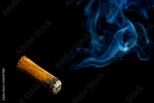 Smoking cuban cigar with blue smoke on an isolated black background. A Havana cigar burns and emits smoke in a dark bar. A rolled cigarette made from tobacco leaves. Burning cigar close up.