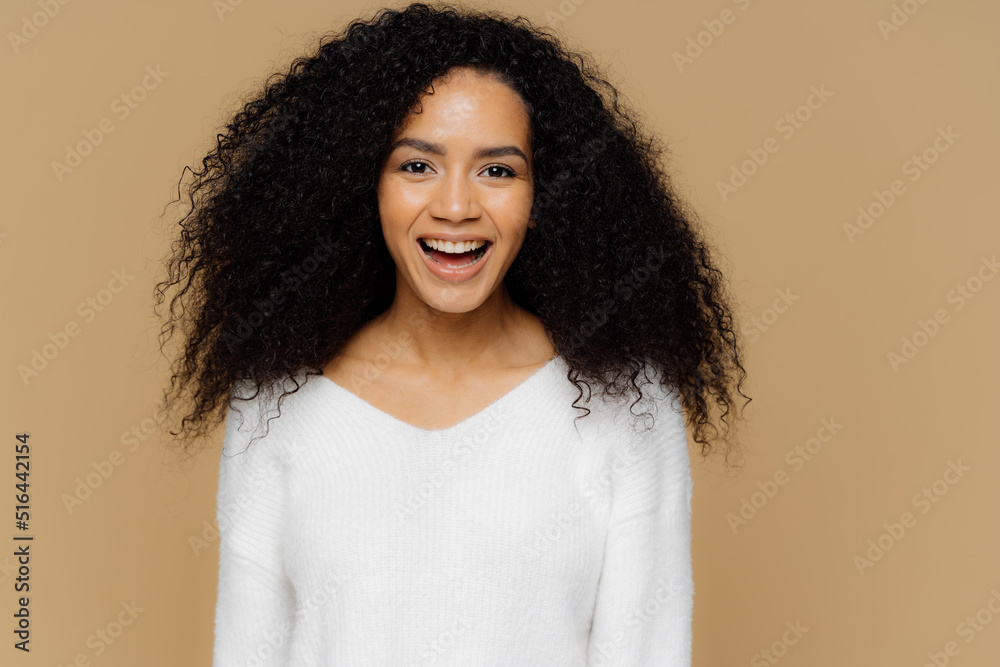 Headshot of healthy positive female model laughs happily, has bushy Afro hair, dressed in white soft jumper, glad to receive compliment from boyfriend, isolated over brown background. Emotions concept