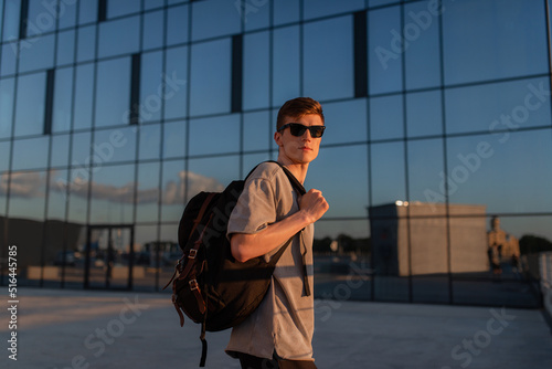 Portrait of handsome tourist hipster man in sunglasses with bag handy, standing guy near the building in downtown. Calm relaxing moment, summer vacation concept