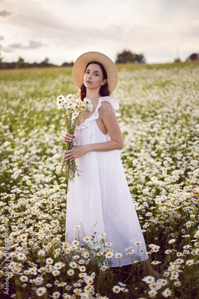 beautiful young brunette woman with long hair in a hat and a white dress is holding a bouquet in her hands standing on a chamomile field at sunset in summer. beautiful young brunette woman with long hair in a hat and a white dress is holding a bouquet in her hands standing on a chamomile field at sunset in summer.