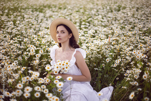 beautiful young brunette woman with long hair in a hat and a white dress is holding a bouquet in her hands standing on a chamomile field at sunset in summer.