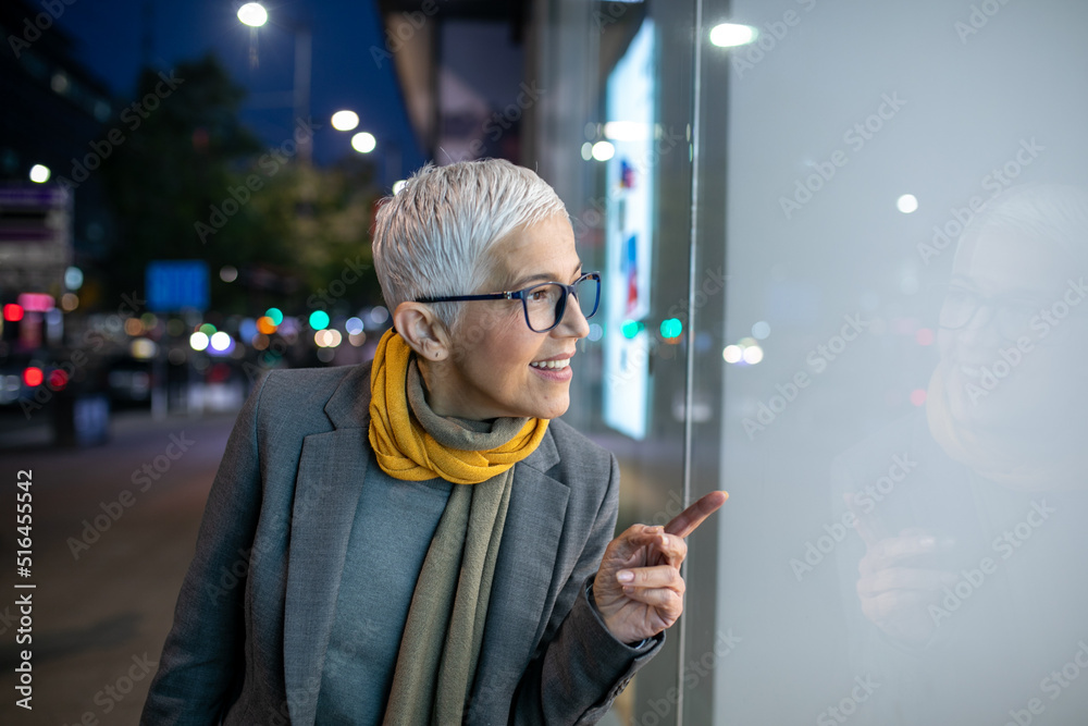 Smiling mature senior woman with short gray hair and eyeglasses looking in the shop window, night scene in city Smiling mature senior woman with short gray hair and eyeglasses looking in the shop window, night scene in city