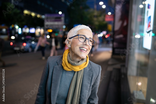 Smiling mature senior woman with short gray hair and eyeglasses walking on street, night scene in city