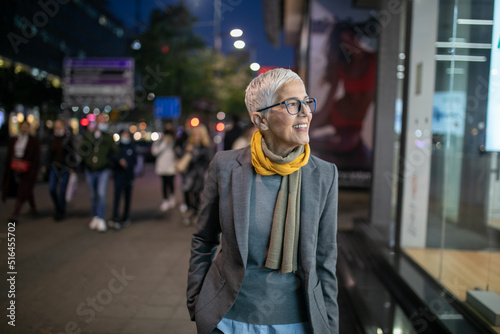 Smiling mature senior woman with short gray hair and eyeglasses walking on street, night scene in city