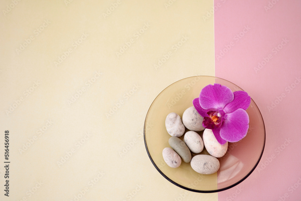 A purple orchid lies in a saucer with sea stones on a yellow and pink background. A purple orchid lies in a saucer with sea stones on a yellow and pink background.