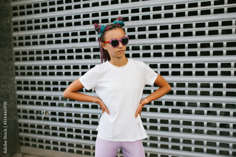 Naklejka premium Portrait of a cute teenage girl with African braids in a white t-shirt and sunglasses stands on a city street. Mock-up for print. T-shirt template.