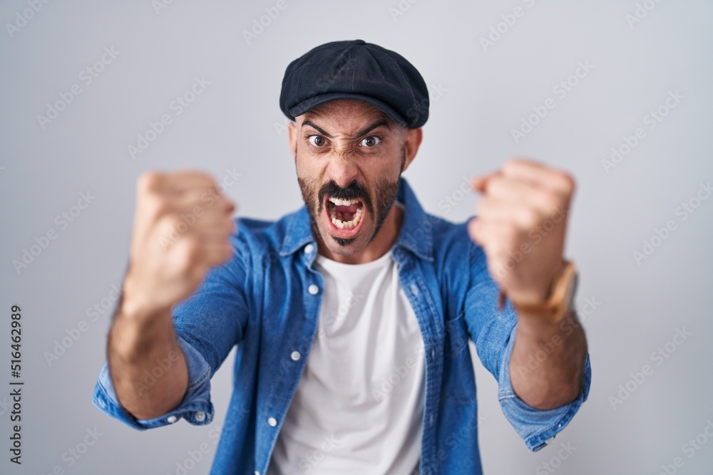 Hispanic man with beard standing over isolated background angry and mad raising fists frustrated and furious while shouting with anger. rage and aggressive concept. Hispanic man with beard standing over isolated background angry and mad raising fists frustrated and furious while shouting with anger. rage and aggressive concept.