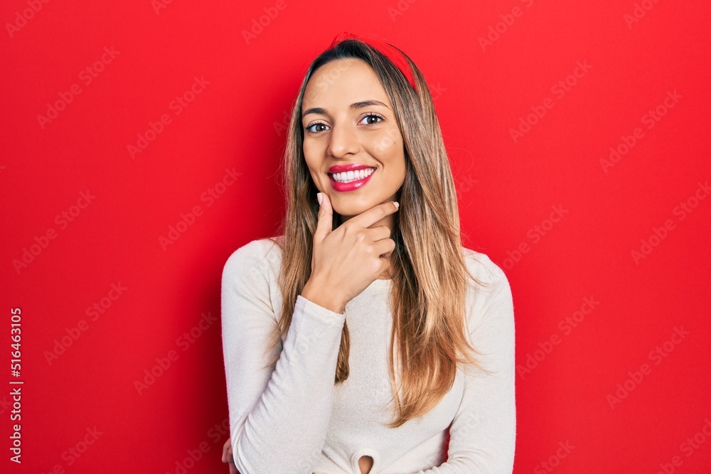 Beautiful hispanic woman wearing red diadem looking confident at the camera smiling with crossed arms and hand raised on chin. thinking positive.