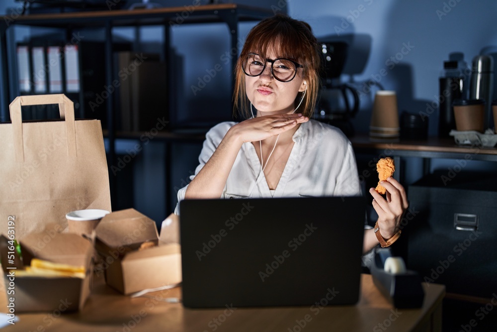 Young beautiful woman working using computer laptop and eating delivery food cutting throat with hand as knife, threaten aggression with furious violence Young beautiful woman working using computer laptop and eating delivery food cutting throat with hand as knife, threaten aggression with furious violence