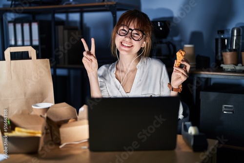 Young beautiful woman working using computer laptop and eating delivery food smiling with happy face winking at the camera doing victory sign with fingers. number two.