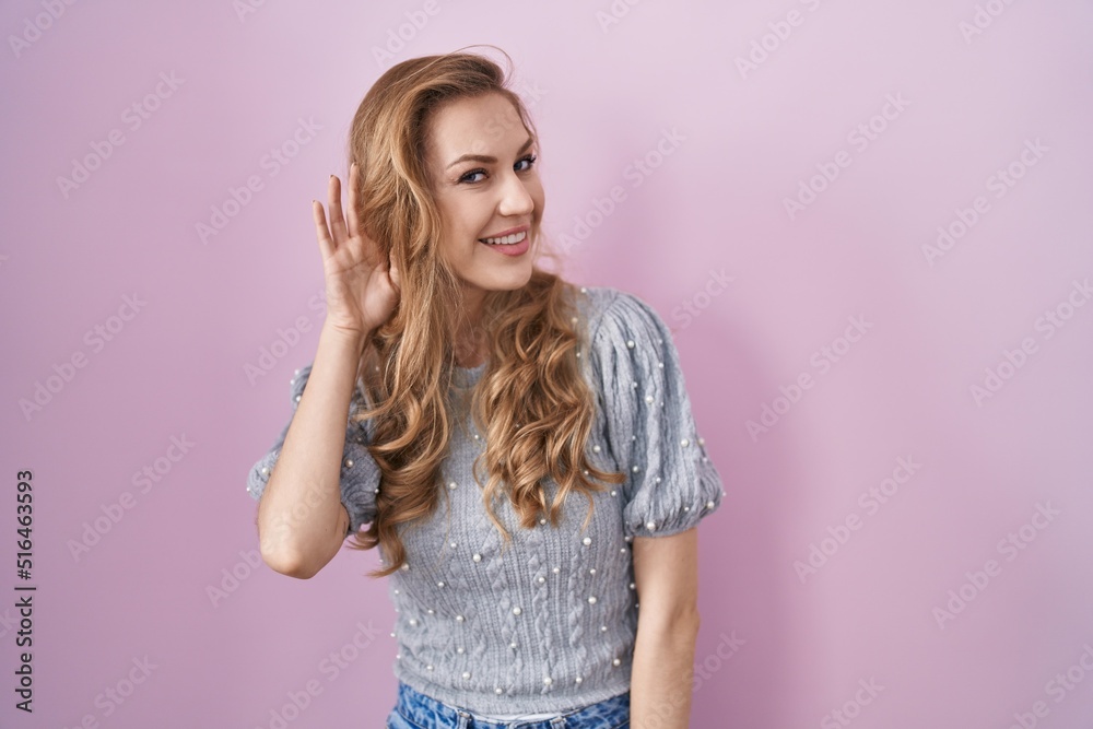 Beautiful blonde woman standing over pink background smiling with hand over ear listening an hearing to rumor or gossip. deafness concept. Beautiful blonde woman standing over pink background smiling with hand over ear listening an hearing to rumor or gossip. deafness concept.