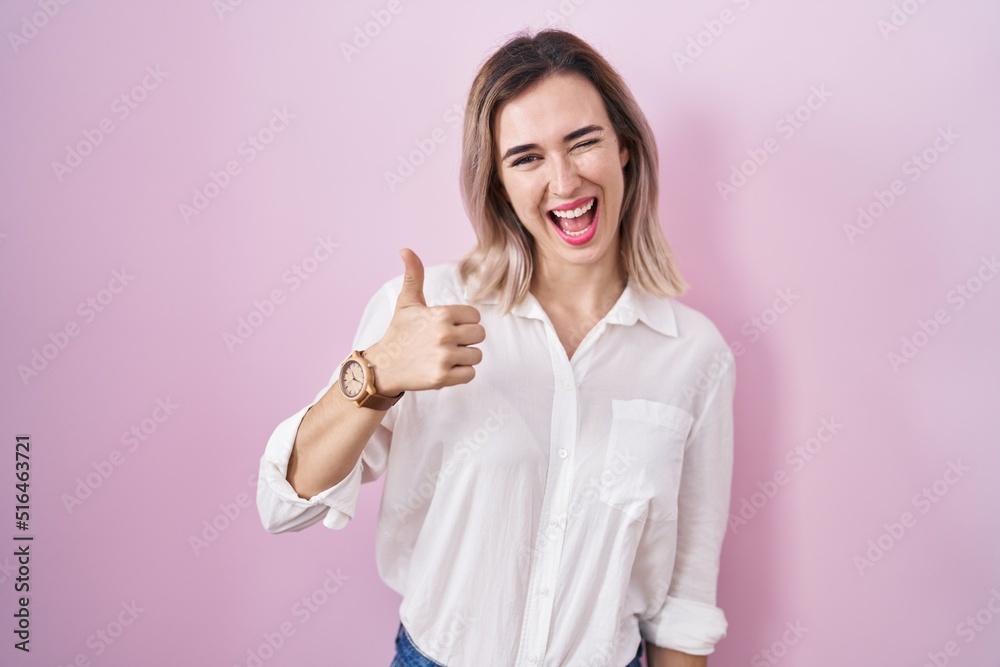 Young beautiful woman standing over pink background doing happy thumbs up gesture with hand. approving expression looking at the camera showing success. Young beautiful woman standing over pink background doing happy thumbs up gesture with hand. approving expression looking at the camera showing success.