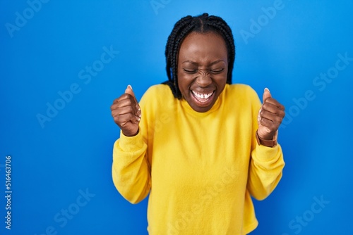 Beautiful black woman standing over blue background excited for success with arms raised and eyes closed celebrating victory smiling. winner concept.