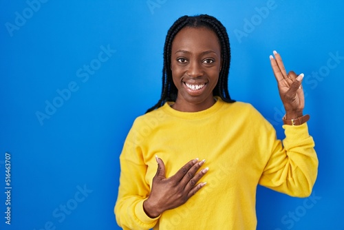 Beautiful black woman standing over blue background smiling swearing with hand on chest and fingers up, making a loyalty promise oath