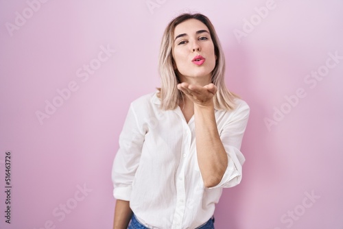 Young beautiful woman standing over pink background looking at the camera blowing a kiss with hand on air being lovely and sexy. love expression.