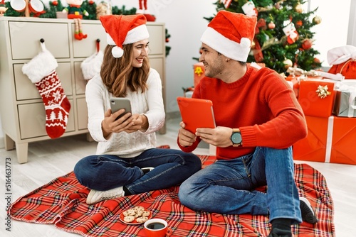 Young hispanic couple wearing christmas hat using touchpad and smartphone sitting on the floor at home.