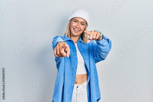 Beautiful blonde woman wearing wool hat pointing to you and the camera with fingers, smiling positive and cheerful