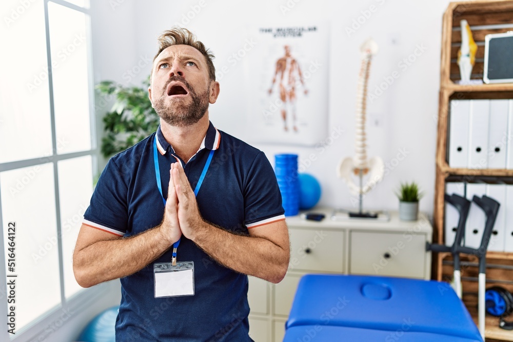 Middle age physiotherapist man working at pain recovery clinic begging and praying with hands together with hope expression on face very emotional and worried. begging. Middle age physiotherapist man working at pain recovery clinic begging and praying with hands together with hope expression on face very emotional and worried. begging.