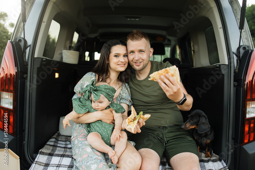 Beautiful married couple with daughter looking at camera while sitting on car trunk with pizza. Celebration, family and happiness concept. © sofiko14