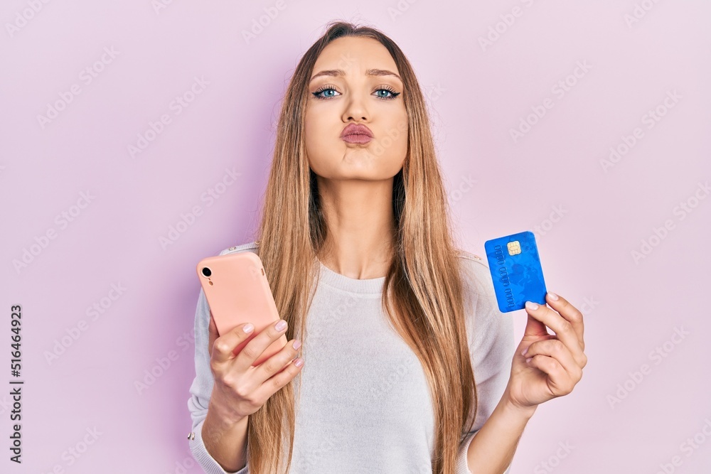 Naklejka premium Young blonde girl holding smartphone and credit card puffing cheeks with funny face. mouth inflated with air, catching air.