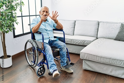 Handsome senior man sitting on wheelchair at the living room showing and pointing up with fingers number six while smiling confident and happy.