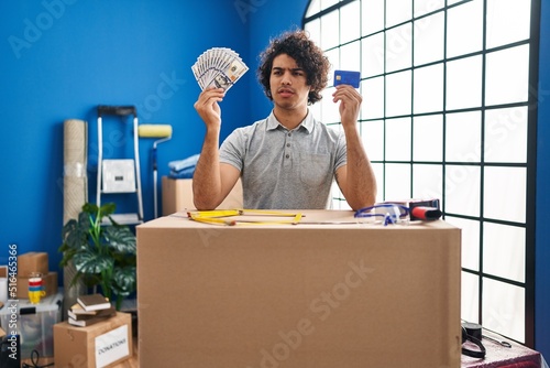 Hispanic man with curly hair moving to a new home holding money and credit card clueless and confused expression. doubt concept. photo