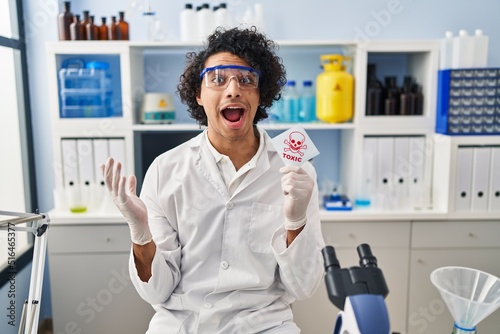 Hispanic man with curly hair working at scientist laboratory holding toxic banner celebrating victory with happy smile and winner expression with raised hands