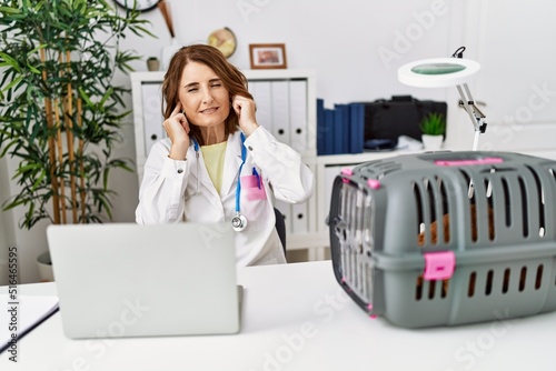 Middle age veterinarian woman working at pet clinic covering ears with fingers with annoyed expression for the noise of loud music. deaf concept.