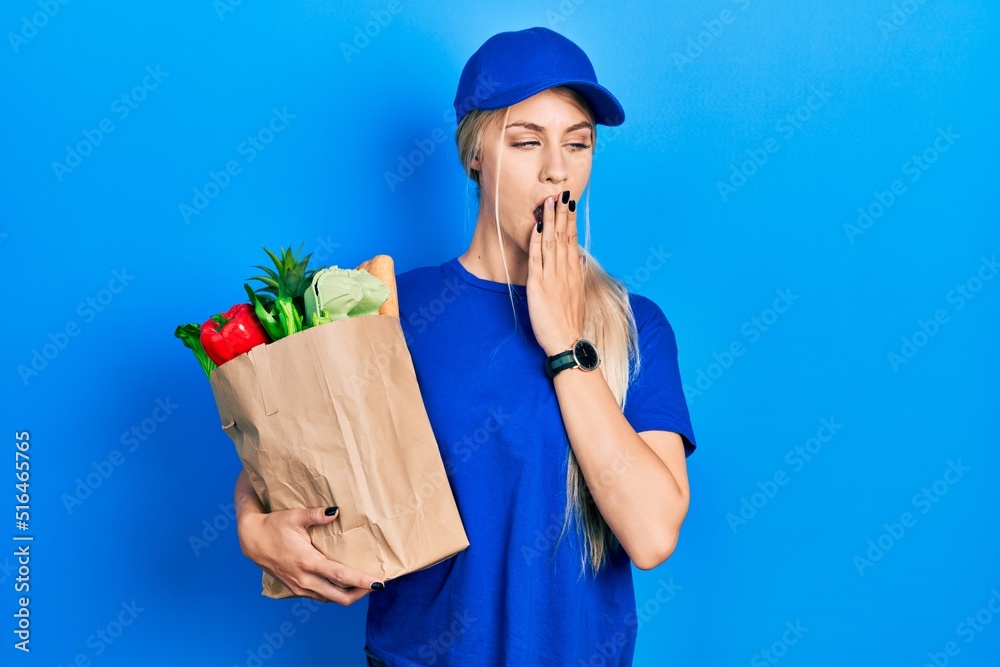 Young caucasian woman wearing courier uniform with groceries from supermarket bored yawning tired covering mouth with hand. restless and sleepiness. Young caucasian woman wearing courier uniform with groceries from supermarket bored yawning tired covering mouth with hand. restless and sleepiness.