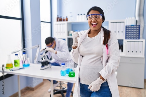Young hispanic woman expecting a baby working at scientist laboratory smiling with happy face looking and pointing to the side with thumb up.