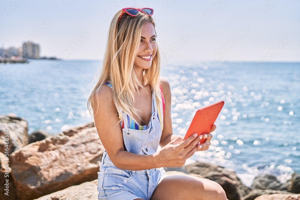 Young blonde girl using touchpad sitting on the rock at the beach.