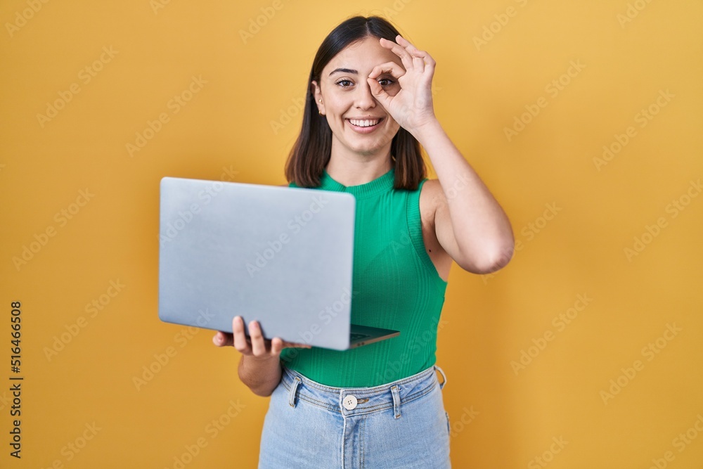Hispanic girl working using computer laptop smiling happy doing ok sign with hand on eye looking through fingers Hispanic girl working using computer laptop smiling happy doing ok sign with hand on eye looking through fingers