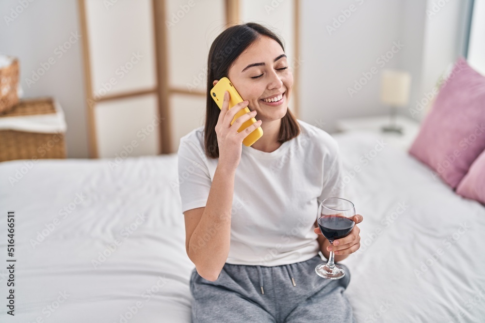 Fototapeta premium Young hispanic woman talking on the smartphone drinking wine at bedroom