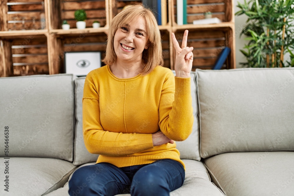 Middle age caucasian woman wearing casual clothes sitting on the sofa at home smiling with happy face winking at the camera doing victory sign. number two. Middle age caucasian woman wearing casual clothes sitting on the sofa at home smiling with happy face winking at the camera doing victory sign. number two.